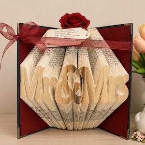 May include: A folded book art sculpture displaying the text Mr & Mrs, featuring a deep red rose on top, a burgundy ribbon tied in a bow, and a small rectangular tag with printed text. The book is set against a dark red interior cover.