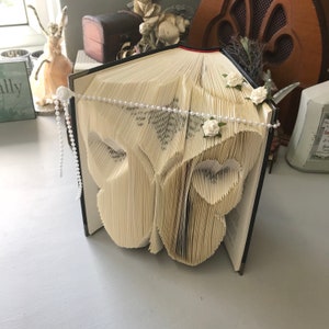 Può includere: Un libro bianco con le pagine piegate a forma di farfalla. Il libro è decorato con una collana di perle e fiori bianchi.