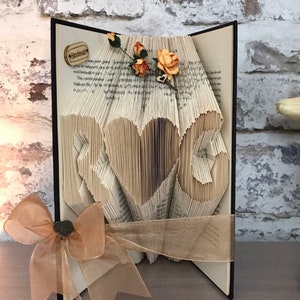 May include: A book art sculpture with the word "LOVE" formed by folding the pages of a book. The sculpture is decorated with a brown ribbon and a small bouquet of orange flowers.