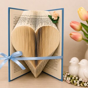 May include: A blue-covered book with pages folded to form a heart shape. A light blue ribbon is tied around the book, and a small peach-colored artificial rose is attached to the top right corner of the pages. The book is open and stands upright.