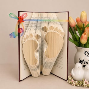 Puede incluir: Una escultura de libro plegado que presenta un par de huellas de pies de bebé creadas doblando las páginas con precisión. Una cinta de colores del arcoíris con un pequeño corazón de madera rojo está atada al lomo. Cubierta marrón oscuro.