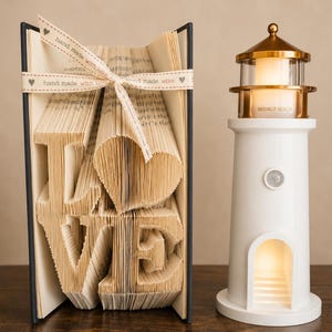 May include: A folded book art sculpture with pages shaped to spell the word LOVE with a heart, tied with a cream ribbon printed with "hand made with love". Beside it stands a white lighthouse-shaped lamp with a gold top and "MOONLIT BEACH" text.