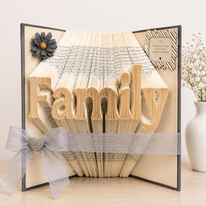 May include: A folded book art sculpture with pages shaped to spell the word Family. The piece features a dark gray fabric flower, a silver ribbon with a heart-shaped rhinestone embellishment, and a small sign with the text: Other things may change us, but we start and end with family.