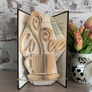 May include: A book art sculpture of a coffee cup with steam rising from it. The sculpture is made from the pages of a book that have been folded and shaped to create the design. The sculpture is displayed on a wooden surface with a vase of flowers in the background.