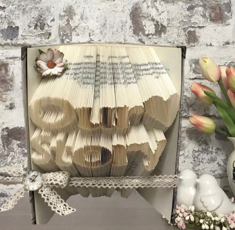 Op de afbeelding: Een boekkunst sculptuur met de woorden "Our Story" gevormd uit gevouwen pagina's. De sculptuur is versierd met een wit kantlint, een klein bloemetje en een vintage-stijl klok.
