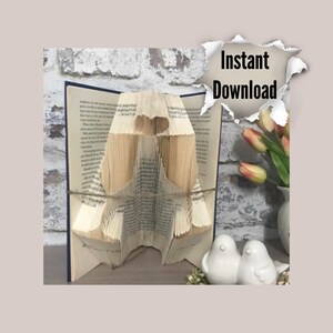 Op de afbeelding: Een boek gevouwen in de vorm van twee vogels met een hart in het midden. De vogels zijn gemaakt van papier en staan op een houten oppervlak. Het boek is open en de pagina's zijn gevouwen in de vorm van de vogels. De vogels zijn wit en het hart is rood. Het boek ligt op een houten oppervlak met een bakstenen muur op de achtergrond.