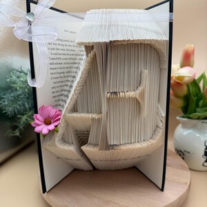 Könnte beinhalten: Eine Buchkunstskulptur des Buchstabens "A", die durch Falten der Seiten eines Buches entstanden ist. Die Skulptur besteht aus einem Taschenbuch mit schwarzem Einband. Das Buch ist geöffnet und die Seiten sind zu einer dreidimensionalen Form gefaltet. Die Skulptur steht auf einer Holzoberfläche. Auf der linken Seite der Skulptur befindet sich eine rosa Blume.