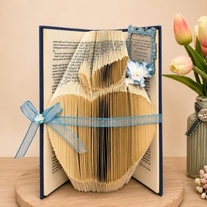 May include: A folded book art sculpture shaped like a heart, featuring a blue ribbon tied around the center. A small blue decorative frame with text is attached to the top right corner, and the book is displayed with a bouquet of pink and white tulips.