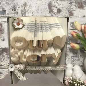 Op de afbeelding: Een boekkunst sculptuur met de woorden "Our Story" gevormd uit gevouwen pagina's. De sculptuur is versierd met een wit kantlint, een klein bloemetje en een vintage-stijl klok.