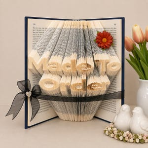 Peut inclure: Un livre aux pages pliées formant le texte Made To Order, avec une couverture bleu foncé, un ruban noir transparent orné d'un petit strass en forme de cœur et une fleur orange foncé fixée sur le coin supérieur droit des pages pliées.