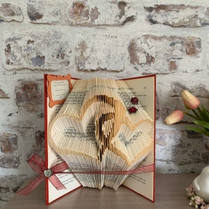 May include: A handmade book art piece with a heart-shaped design, created by folding the pages of a book. The open book is red with a burgundy ribbon and a decorative embellishment. The text on the book reads "To Amy and Rob..." and "Happy 27th Anniversary".