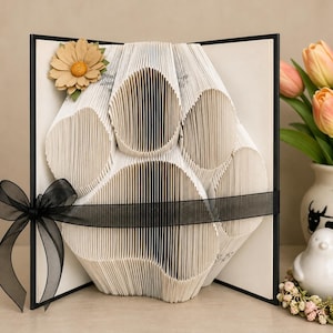 Peut inclure: Une sculpture en livre plié en forme d'empreinte de patte, composée de pages blanches aux plis complexes. Un ruban noir transparent est noué horizontalement autour du centre, et une fleur en papier beige est fixée dans le coin supérieur gauche.