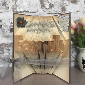 Può includere: Una scultura di arte di libro con la parola "famiglia" piegata nelle pagine. Il libro è decorato con un piccolo fiore e un nastro. Il libro è seduto su un tavolo con un vaso di fiori sullo sfondo.