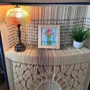 May include: A unique book art piece featuring a miniature lamp with a textured shade, a small framed painting of tulips, and a potted plant. The book pages are intricately folded and cut to create a decorative shelf and backdrop.