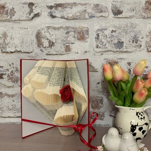 May include: A red rose crafted from the pages of a book, with the pages folded into a heart shape. The book is open and standing on a table, with a red ribbon tied around it. There are tulips in a vase and two white ceramic birds on the table.