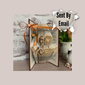 May include: A book art sculpture with the words "Be Brave" folded into the pages. The book is open and standing upright on a wooden surface. The sculpture is decorated with a brown ribbon and orange flowers.