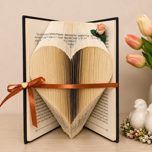 May include: A book with pages folded to form a heart shape, featuring a brown satin ribbon tied around the centre with a small gold heart charm. A miniature pink rose is attached to the top right corner of the folded pages, which are open.