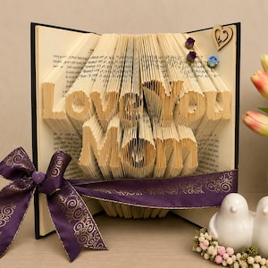 May include: A folded book art sculpture with pages shaped to spell out the words Love You Mom. The book is decorated with a purple ribbon featuring a gold swirl pattern, a small purple heart, two tiny fabric flowers, and a wooden heart cutout.