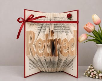 May include: A folded book art sculpture with pages shaped to spell the word Retired. The book is framed by a dark red cover and accented with a thin dark red ribbon tied in a bow on the left side, plus a small red fabric rose at the top right.