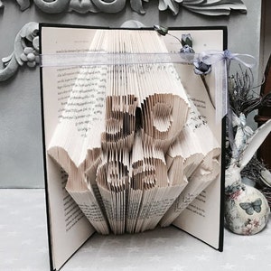 May include: A book art sculpture, folded into the shape of the word "LOVE", with a white ribbon and a blue flower on the cover. The book is open and standing on a white surface.