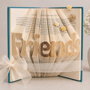 Pode incluir: Uma escultura de livro dobrado com as páginas meticulosamente vincadas para formar a palavra Friends. O livro apresenta uma capa azul-petróleo, uma fita branca transparente atada num laço na frente e pequenas flores decorativas de tecido amarelo.