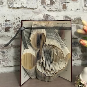 May include: A book art sculpture of a heart made from the pages of a book. The book is bound in red and has a black ribbon tied around it. The sculpture is on a table with a white brick wall in the background.