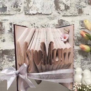 Può includere: Una scultura di arte di libro con le parole "My Mum" formate dalle pagine. Il libro è decorato con un nastro bianco e un piccolo fiore rosa.
