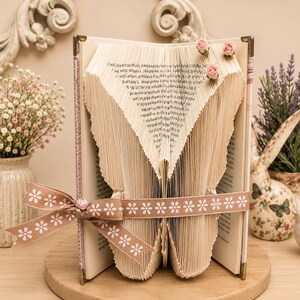 Può includere: Una scultura di libro con pagine piegate a forma di cuore, decorata con tre piccole rose di tessuto rosa sulla parte superiore. Un nastro marrone chiaro con un motivo a margherite bianche è legato al centro, fissato con una piccola perlina a forma di cuore rosa.