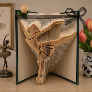 Può includere: Una scultura di libro con pagine piegate che formano la sagoma di una ballerina in posa. Il libro è decorato con una singola rosa artificiale color pesca nell'angolo in alto a sinistra e un nastro nero trasparente annodato sul lato destro.
