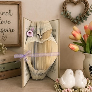 May include: A book art sculpture featuring pages folded into a heart shape, accented with a small purple calla lily and a light purple ribbon tied around the center. The sculpture stands upright, displaying the text of the original book pages.