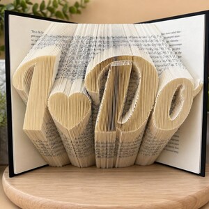 May include: A book sculpture featuring folded pages that form the text "I Do" with a heart symbol between the words. The book has a black cover and is displayed in an open position, showcasing the intricate paper folding technique used to create the letters.