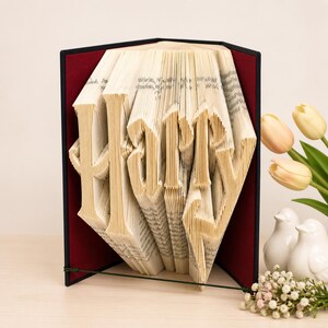 Könnte beinhalten: Eine Buchfaltkunst-Skulptur, die das Wort Harry bildet, indem die Seiten eines Buches präzise gefaltet wurden. Die Skulptur befindet sich vor einem dunkelroten Hintergrund in einem schwarzen Rahmen und zeigt die komplexe Falttechnik.