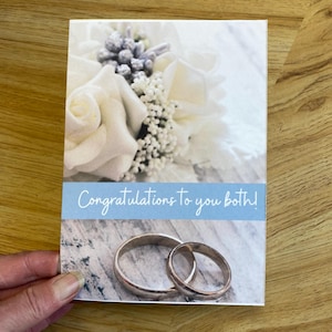 May include: A wedding greeting card with a floral design and two wedding rings. The card features a light blue banner with the text "Congratulations to you both!" The background has a wood-like texture.