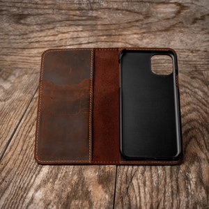 May include: A brown leather phone case with a flap that opens to reveal a card holder and a phone slot. The case is closed and lying on a wooden surface.