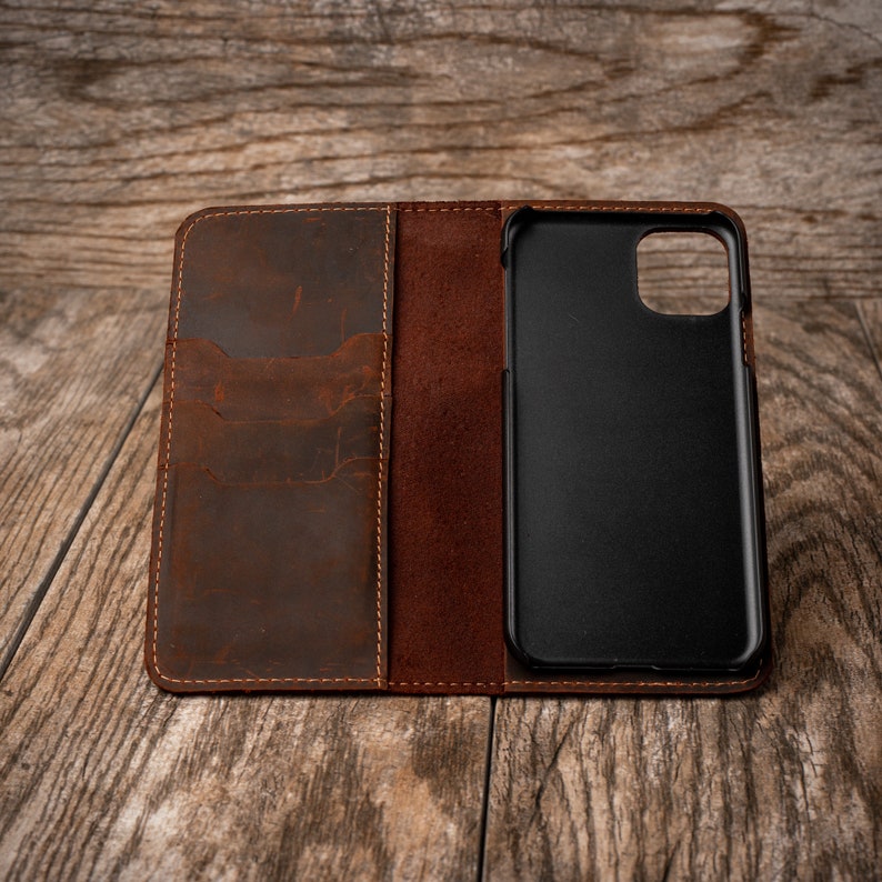 May include: A brown leather phone case with a flap that opens to reveal a card holder and a phone slot. The case is closed and lying on a wooden surface.