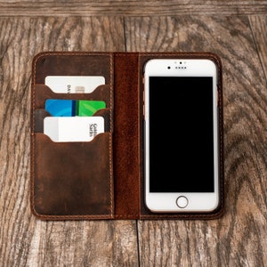 May include: A brown leather phone case with slots for credit cards. The case has a stitched edge and a phone slot with a white phone inside.