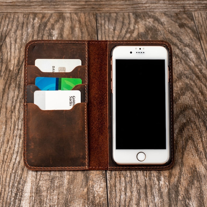 May include: A brown leather phone case with slots for credit cards. The case has a stitched edge and a phone slot with a white phone inside.