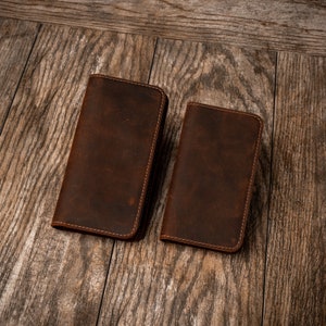 May include: Two brown leather wallets with stitched edges. The wallets are laid out on a wooden surface.