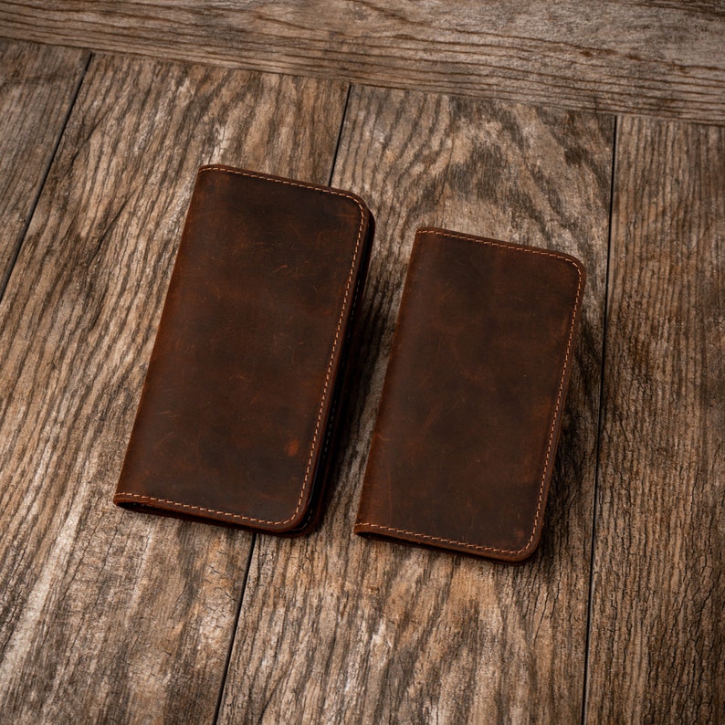 May include: Two brown leather wallets with stitched edges. The wallets are laid out on a wooden surface.
