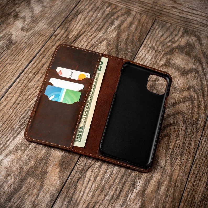 May include: A brown leather phone case with a built-in wallet. The case has slots for credit cards and a pocket for cash. The phone case is open and a black phone is visible inside.