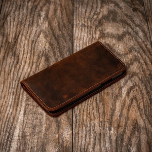 May include: A brown leather phone case with stitching around the edges. The case is lying on a wooden surface.