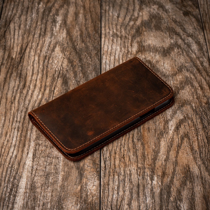 May include: A brown leather phone case with stitching around the edges. The case is lying on a wooden surface.