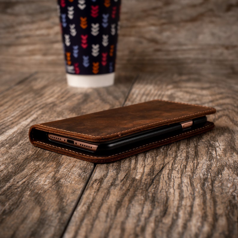 May include: A brown leather phone case with a black phone inside. The case is lying on a wooden surface.