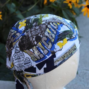 May include: A white scrub cap featuring a collage of University of Michigan imagery, including the word Michigan in large block letters, stadium graphics, and the letter M in navy blue and yellow, with tie-back straps and a patterned fabric design.
