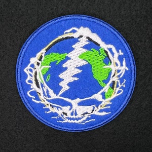 Peut inclure: Un patch brodé bleu et vert avec un éclair et une carte du monde. Le patch est entouré d'un contour blanc.