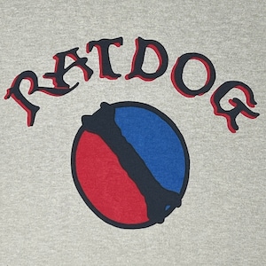 May include: A light gray t-shirt featuring the word "RATDOG" in a red, arched font with a black outline. Below the text is a circular graphic divided into red and blue sections with a bone design.