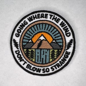 Peut inclure: Écusson brodé circulaire avec un motif de montagne et de soleil dans les tons marron, orange et bleu. Le texte "GOING WHERE THE WIND DON'T BLOW SO STRANGE" est brodé en noir sur le bord. Idéal pour les vêtements ou les sacs.