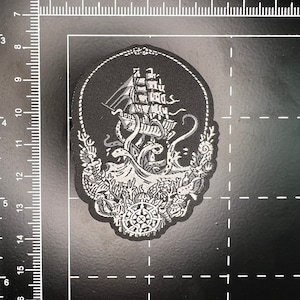 Puede incluir: Un parche bordado en blanco y negro que representa un barco siendo atacado por un pulpo gigante. El barco está rodeado de imágenes náuticas detalladas, incluyendo una brújula, una cuerda y algas marinas.