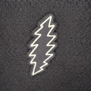 Peut inclure: Un patch noir avec un éclair blanc. L'éclair est stylisé et a un bord dentelé.
