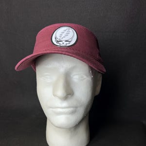May include: A burgundy visor featuring a white and black graphic of a skull with a lightning bolt design. The visor is displayed on a white mannequin head against a dark background. The visor has a curved brim.